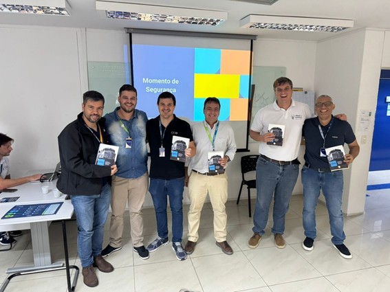 Grupo de seis homens um ao lado do outro segurando um livro. Ao fundo um telão.