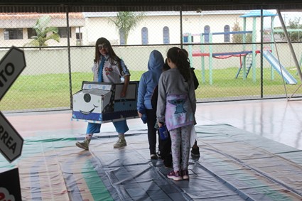 Crianças em fila acompanham uma mulher que segura um modelo de caminhão, em área coberta com piso colorido e playground ao fundo.