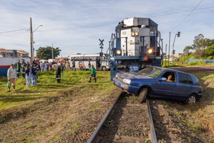 Carro azul está preso nos trilhos de trem, com a frente sobre a linha férrea, enquanto um trem se aproxima e pessoas observam a cena ao redor.