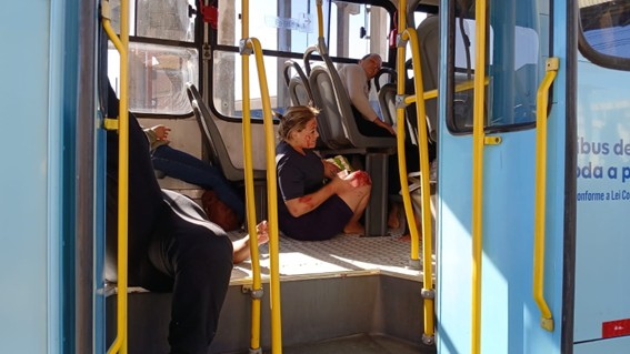 Pessoas sentadas e deitadas dentro de um ônibus azul com portas abertas.
