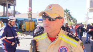 Homem com capacete e uniforme de bombeiro, olhando para a câmera em área externa.