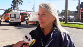 Mulher sendo entrevistada, com microfone e ambulâncias ao fundo.