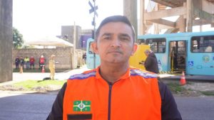 Homem com colete laranja de resgate, com ônibus e pessoas ao fundo.