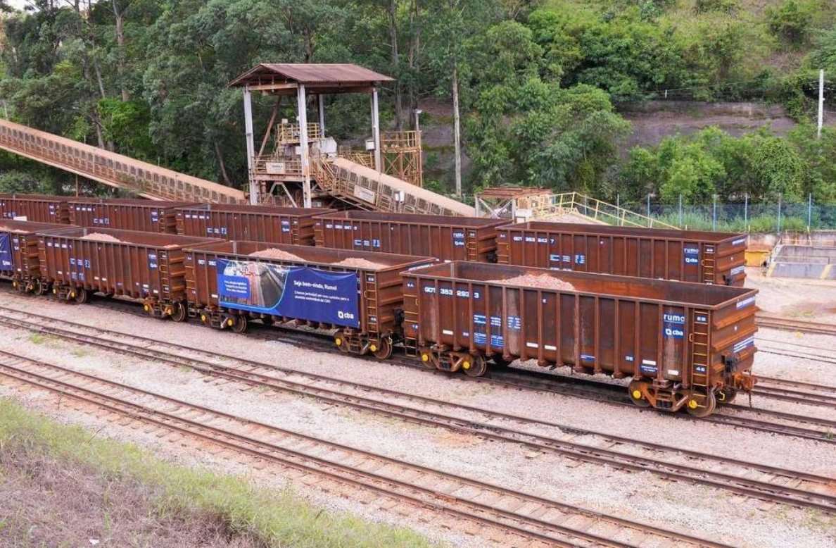 Fileira de vagões de trem marrons, carregados com material a granel, em trilhos paralelos. Ao fundo, uma estrutura metálica com uma passarela elevada e vegetação densa. A cena sugere uma área industrial ou de transporte de carga.