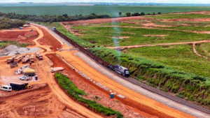 Uma vista aérea de uma ferrovia em construção, com um trem em movimento. Ao lado da ferrovia, há áreas de terraplanagem e vegetação. O céu é nublado.