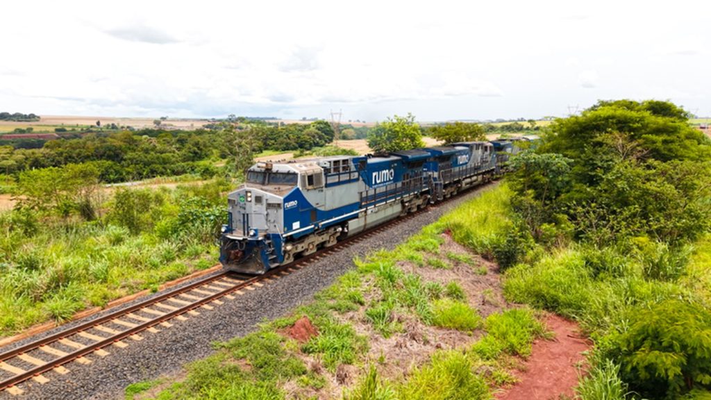 Uma locomotiva azul e cinza puxando vagões em uma ferrovia. A cena é em um ambiente rural, com vegetação exuberante ao lado dos trilhos. O céu é claro.