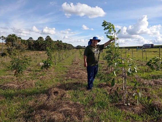 Um homem sorrindo, usando chapéu, em um campo com fileiras de árvores jovens. Ele está tocando em uma das árvores. O céu é azul com algumas nuvens.