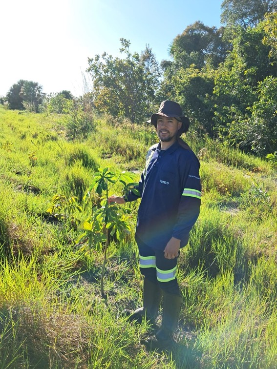 Um homem em um macacão azul escuro, com detalhes refletivos, em um campo gramado. Ele está em pé ao lado de uma muda de árvore, com a mão sobre ela. Ao fundo, vegetação e céu ensolarado.