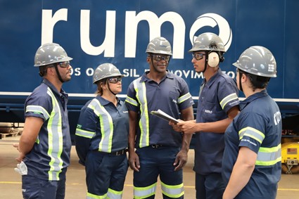 Grupo de cinco pessoas usando uniforme da Rumo e equipamentos de segurança. Ao fundo, um vagão escrito Rumo.