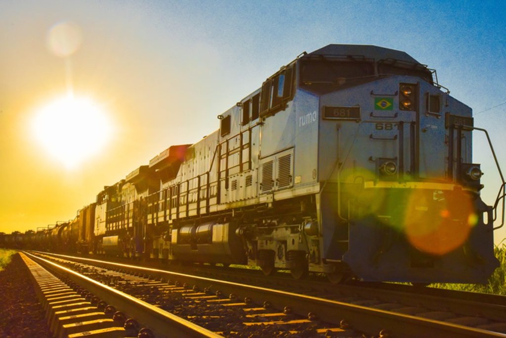 Trem de carga com múltiplas locomotivas brancas e amarelas em trilho ao amanhecer, com sol brilhante ao fundo iluminando a composição e refletindo nos trilhos.