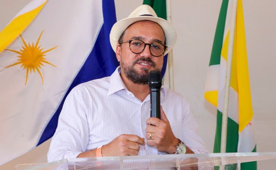 Homem usando óculos e chapéu branco discursa em microfone durante evento, com bandeiras brasileiras e municipais ao fundo, representando momento importante da solenidade.