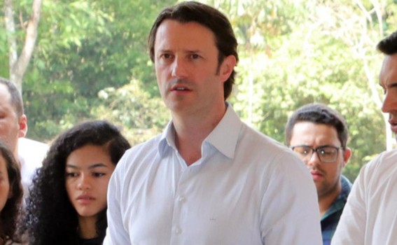 homem com camisa social clara discursando, com pequeno grupo de pessoas ao fundo e vegetação verde desfocada.