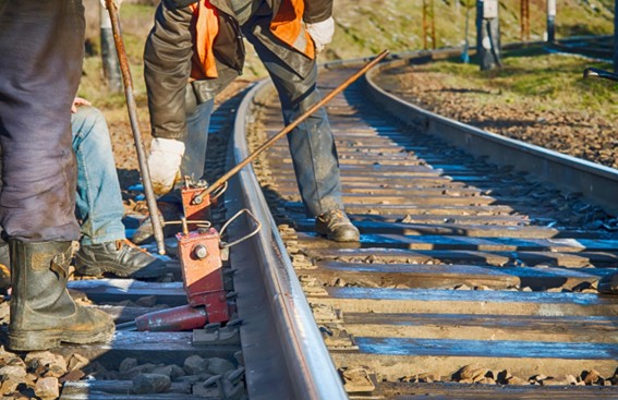 Operários com botas e equipamentos de proteção trabalhando na manutenção de trilhos de ferrovia, utilizando ferramentas