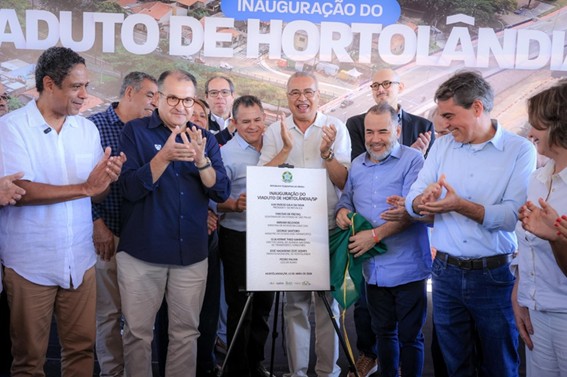 Grupo de autoridades e convidados sorridentes e aplaudindo ao redor de uma placa oficial durante a cerimônia de inauguração do Viaduto de Hortolândia. Ao fundo, há um painel com o nome do evento.