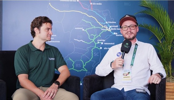 Dois homens sentados em poltronas durante entrevista, um deles segurando um microfone, com mapa da malha ferroviária ao fundo.